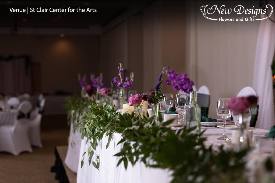 A stunning head table of trailing green garland, and bud vases of various heights sporting purple flowers in various textures and shades.